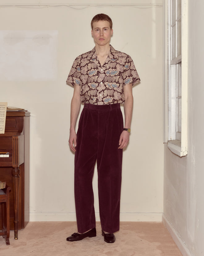 A person stands indoors by a wooden piano, wearing a floral shirt and Corduroy McNab Trousers in dark purple with a high-rise, wide-leg silhouette.