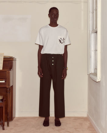 A person stands indoors by a piano and window, wearing a white T-shirt and Astor Pajama Pants in black, featuring a heavyweight wool fabric and drawstring waist.