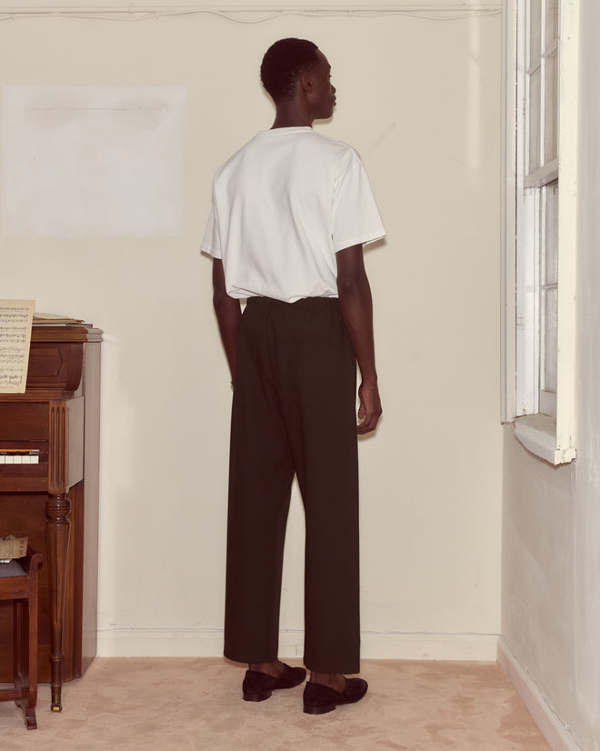 A person stands indoors facing a wall, wearing a white t-shirt, black Astor Pajama Pants with a drawstring waist, and slippers.