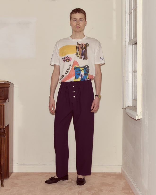 A man stands indoors wearing a graphic t-shirt, the Astor Pajama Pants in purple with a drawstring waist and buttons, and black shoes.