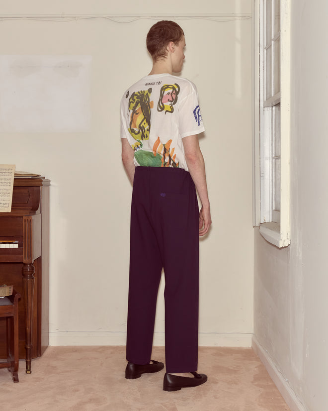 A person stands indoors facing a wall, wearing a graphic t-shirt, Astor Pajama Pants - Purple with a drawstring waist, and black shoes.