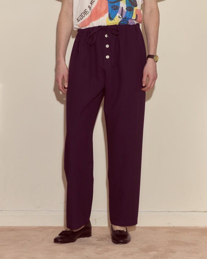 Person wears Astor Pajama Pants - Purple, paired with a graphic t-shirt and black loafers while standing indoors.