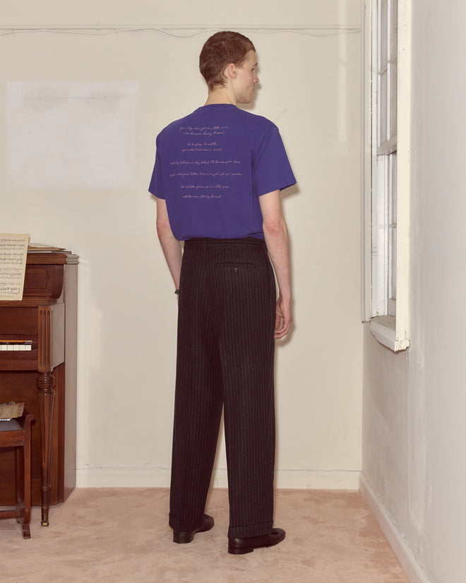 A person stands indoors facing away, wearing a blue shirt and Waldorf Pinstripe Trousers with side buckle waist adjusters near a piano and window.