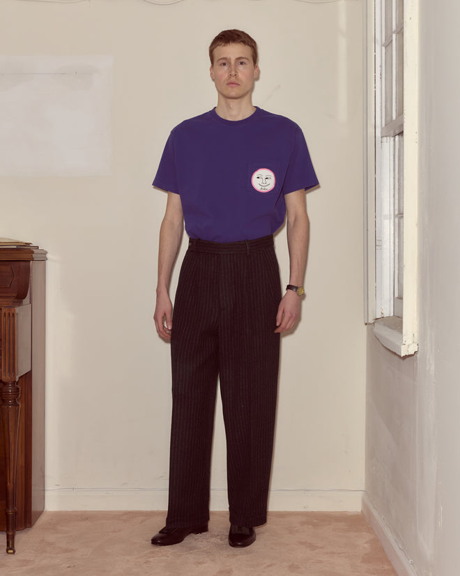 A man stands in a plain room next to a window, facing the camera, wearing a purple T-shirt and Waldorf Pinstripe Trousers.