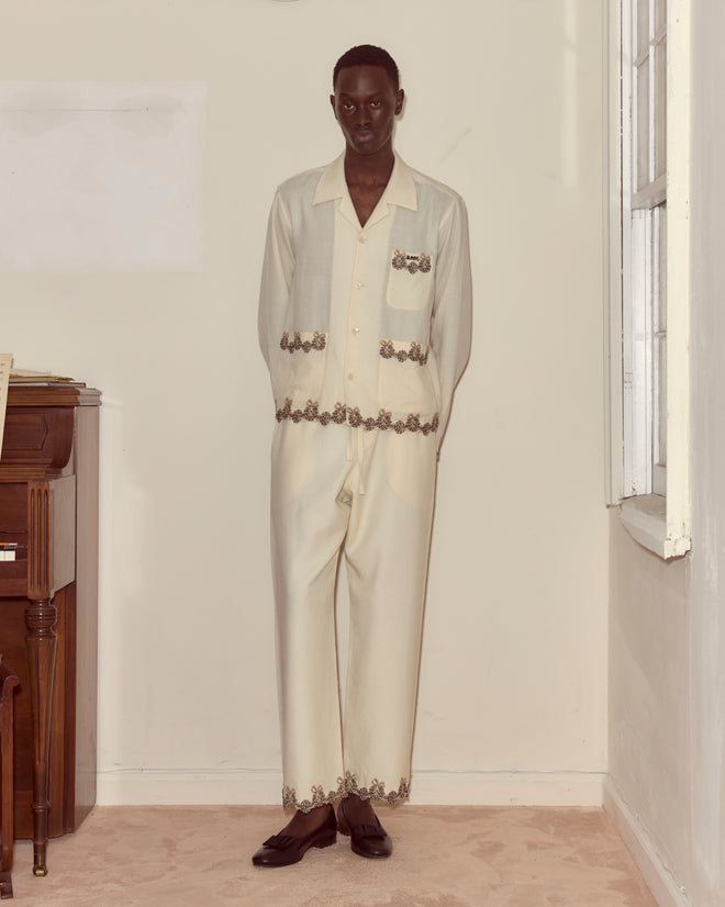 A person stands indoors wearing Laurelist Pajama Pants in cream, featuring embroidered and vintage passementerie details, paired with black shoes and hands behind their back.