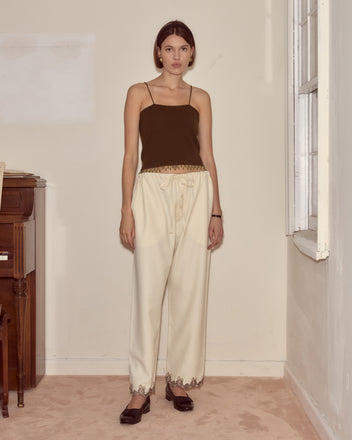 A woman in a brown tank top and Laurelist Pajama Pants in cream stands indoors on a beige carpet near a window and piano, her outfit accented with vintage passementerie for timeless elegance.