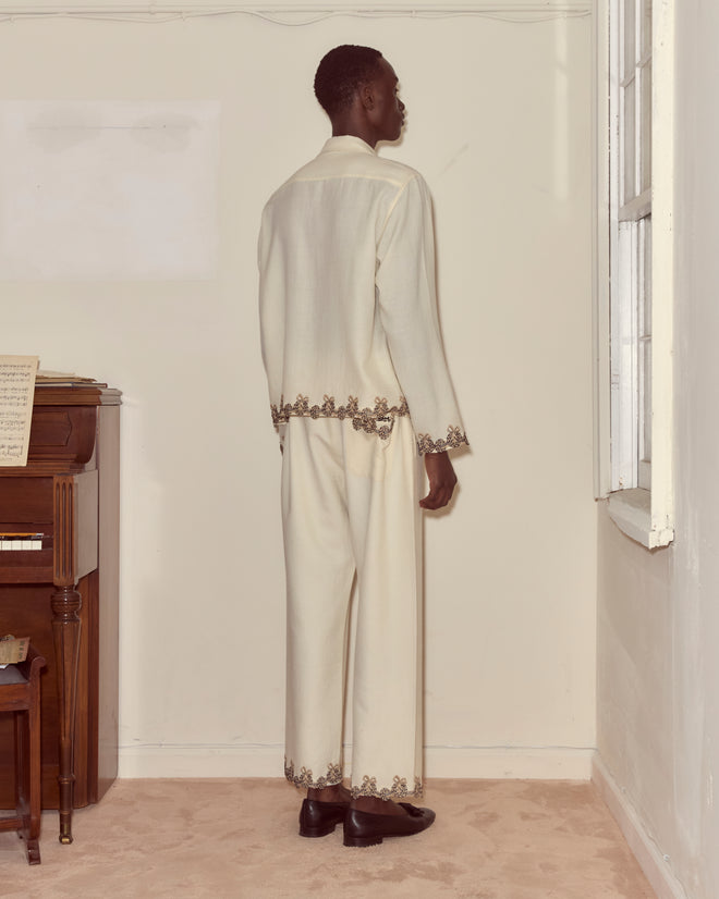 A person wearing Laurelist Pajama Pants in cream stands by a piano and window, where soft flannel drapes and vintage passementerie add texture to the softly lit room.