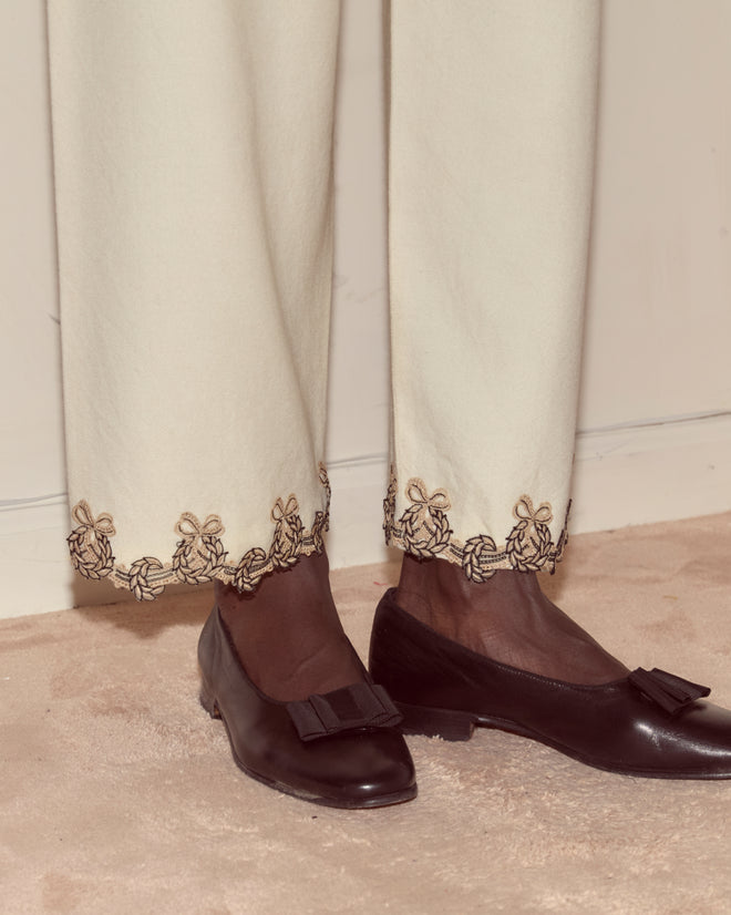 Person wearing Laurelist Pajama Pants in cream with vintage passementerie embroidered hem and black bow loafers, standing on a beige carpet.