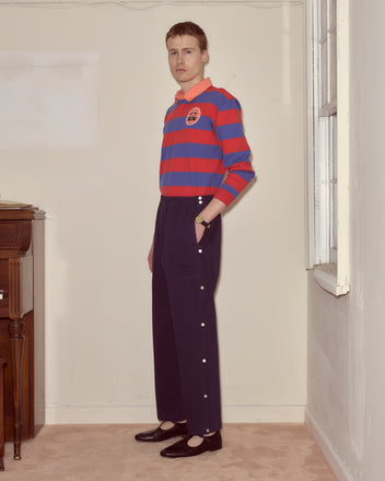 A man in a striped rugby shirt and Twill Tear-Away Track Pants - Navy stands indoors by a window, looking at the camera.