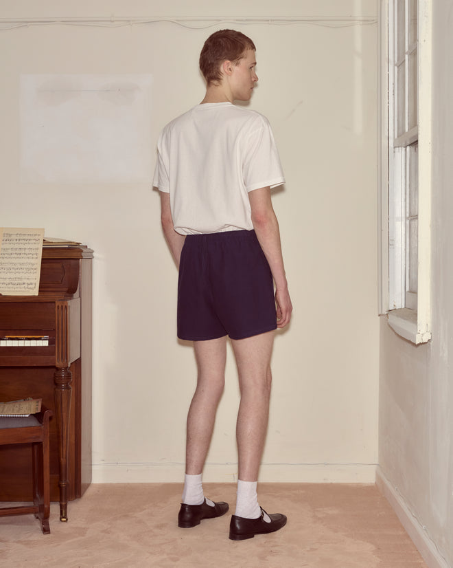 Wearing Field Shorts - Navy, made from heavyweight cotton twill, a person in a white shirt stands facing a window in a sparsely furnished room, the fabric lending structure to the relaxed atmosphere.