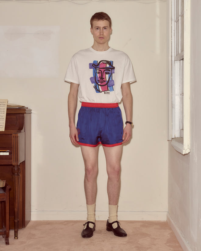 A man in a graphic t-shirt, Heart Deco Gym Shorts - Blue, socks, and Mary Jane shoes stands by a window in a sparse room, channeling a 1970s silhouette.