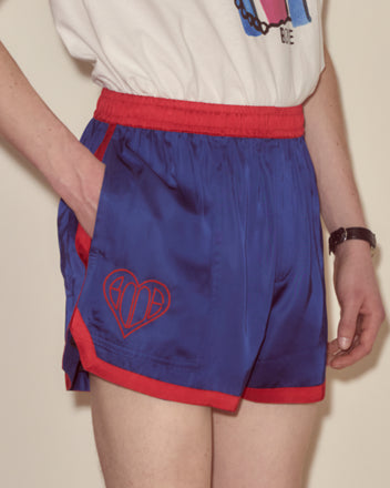 A person wearing Heart Deco Gym Shorts - Blue, featuring red trim in a 1970s silhouette with heart embroidery, paired with a white graphic tee.