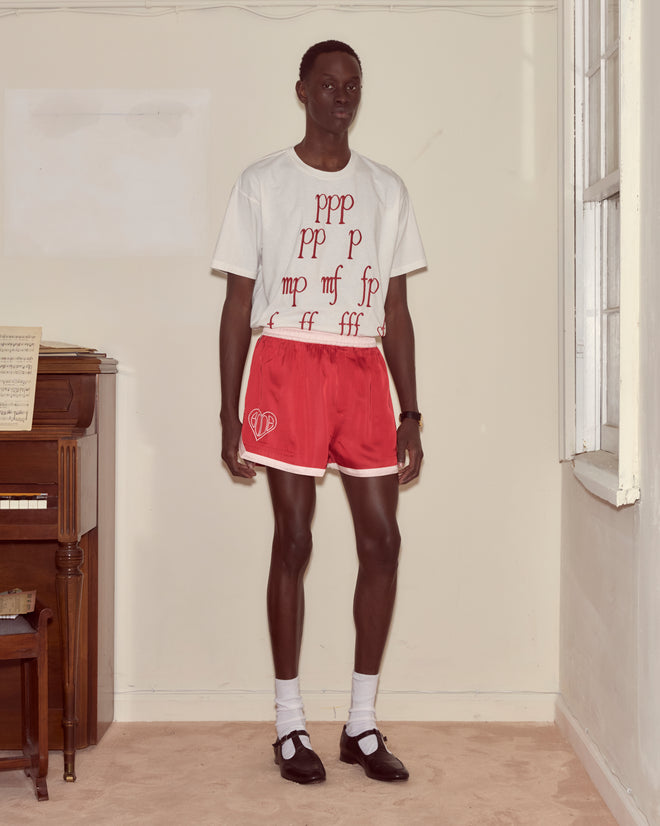 A person stands indoors wearing a white graphic t-shirt and Heart Deco Gym Shorts - Red, paired with socks and black shoes for a 1970s-inspired look.