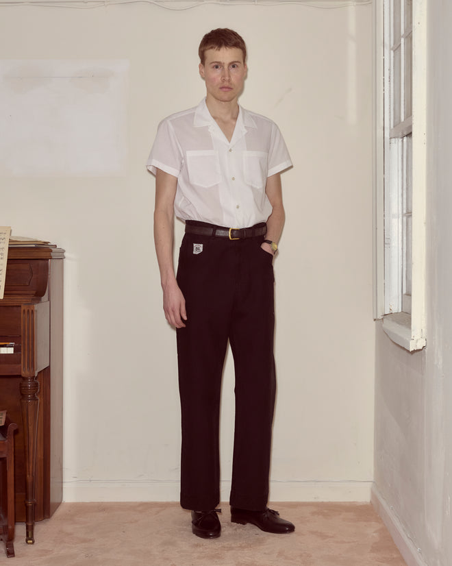 A person stands indoors in a white European smock and Embroidered Denim Knolly Brook Trousers - Black, beside a piano and a window.