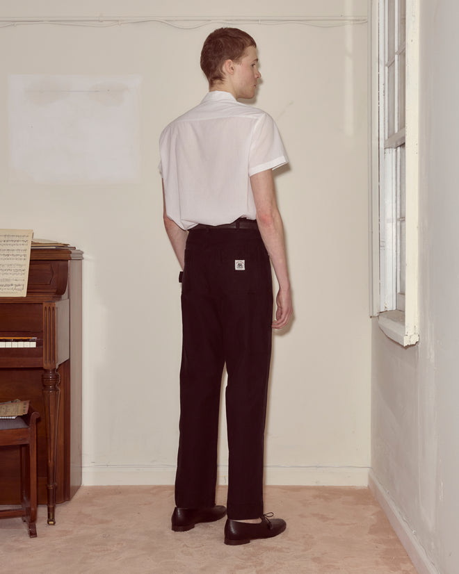 Wearing a 100% cotton white shirt and black Embroidered Denim Knolly Brook Trousers, a person stands facing a corner near a piano and window.