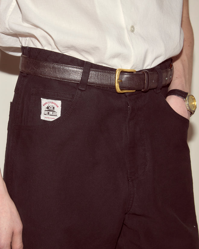 Person wearing a white shirt, Embroidered Denim Knolly Brook Trousers - Black, a brown belt, and a wristwatch with a gold face.