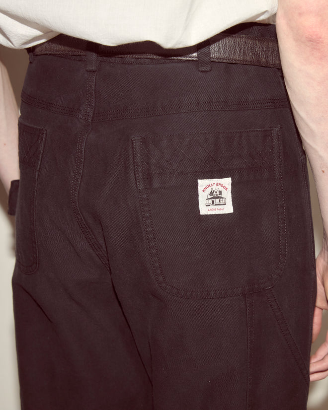 A person wears black Embroidered Denim Knolly Brook Trousers with a visible “WALLY BROWN” back pocket label, made from 100% cotton, and a white shirt tucked in.