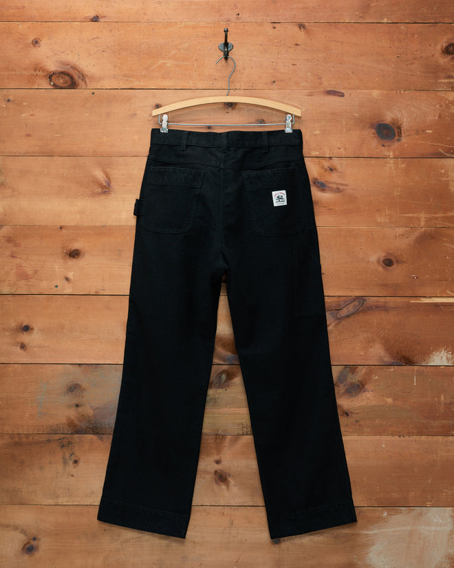 Embroidered Denim Knolly Brook Trousers in black hanging on a wooden hanger against a rustic wood wall, viewed from the back—a classic Bode piece that blends timeless style with everyday versatility.