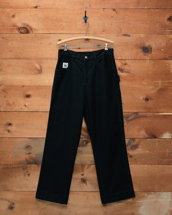 Embroidered Denim Knolly Brook Trousers - Black hang neatly on a hanger against a rustic wooden wall, highlighting their timeless appeal. These black embroidered trousers seamlessly blend sophistication and craftsmanship, making them an ideal addition to the modern wardrobe.