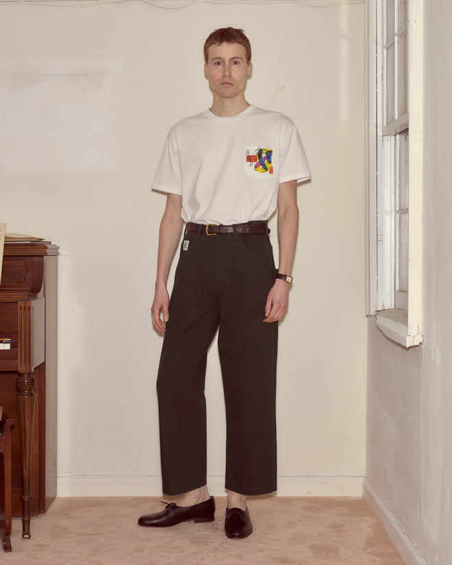 A person stands indoors by a piano wearing a white graphic t-shirt, Twill Knolly Brook Trousers in dark green with a side pocket, and loafers.