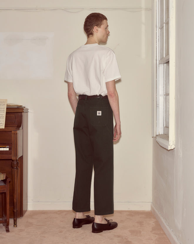 A person in a white t-shirt and Twill Knolly Brook Trousers - Dark Green with a side pocket stands indoors, facing a wall near a piano and window.