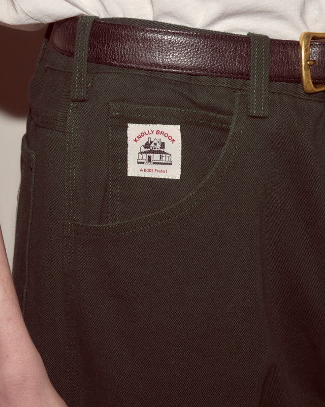 Someone wears Twill Knolly Brook Trousers - Dark Green, paired with a brown belt, featuring the Knolly Brook label above the side pocket.