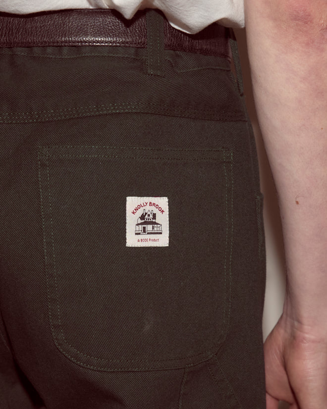 Close-up of a person wearing dark green Twill Knolly Brook Trousers with a white "Muddy Brook" patch on the back pocket.