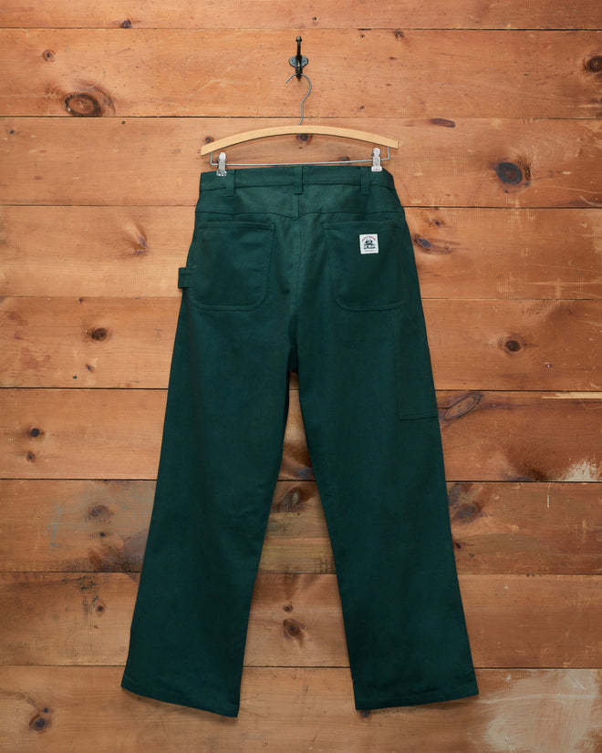 The Twill Knolly Brook Trousers in dark green, featuring back pockets, are shown on a hanger against a wooden wall background.