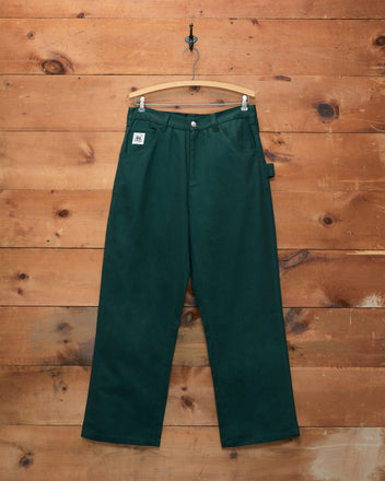 The Twill Knolly Brook Trousers in dark green, made from cotton twill, hang on a hanger against a wooden wall, featuring a tag on the front pocket.