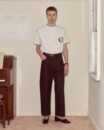 Man wearing a white t-shirt and Waxed Cotton Knolly Brook Trousers in brown stands indoors by a piano and window, looking at the camera.