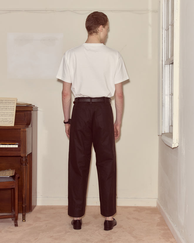 A person stands facing a wall in a simple room, wearing a white t-shirt, Waxed Cotton Knolly Brook Trousers - Brown, and black shoes.