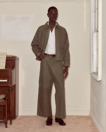 A man in an olive green suit and white shirt stands by a piano and window, his look completed with Waxed Cotton Knolly Brook Trousers in Light Green for a nod to timeless style.