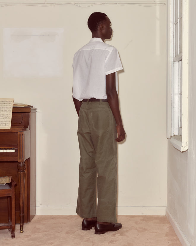 A person in a white shirt and Waxed Cotton Knolly Brook Trousers - Light Green stands facing a corner of a beige room near a piano.