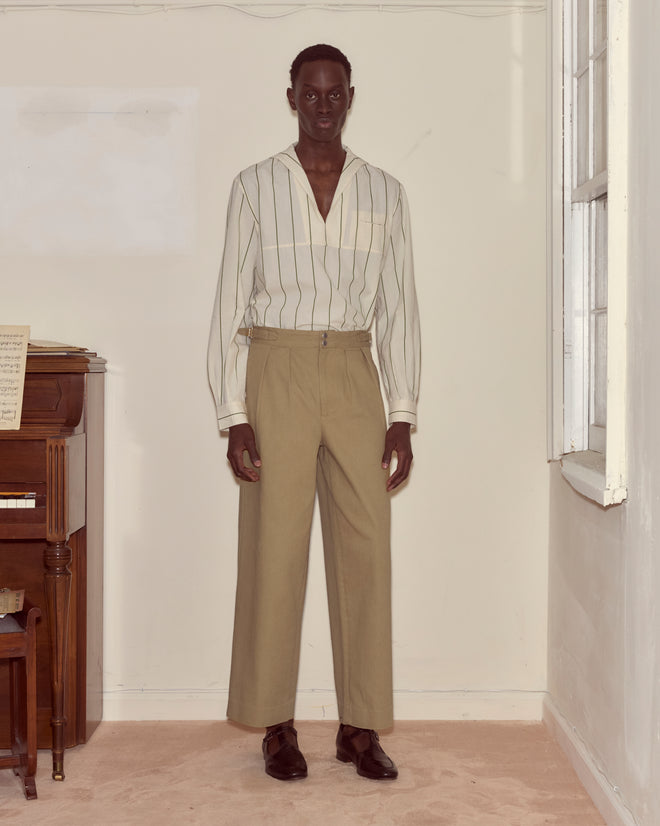 A man stands indoors by a piano and window, wearing a striped shirt and Wide Leg Snap Trousers - Khaki.