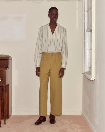 Indoors by a window and wooden table, a person wears a striped shirt with Wide Leg Snap Trousers - Khaki featuring side buckle waist adjusters.