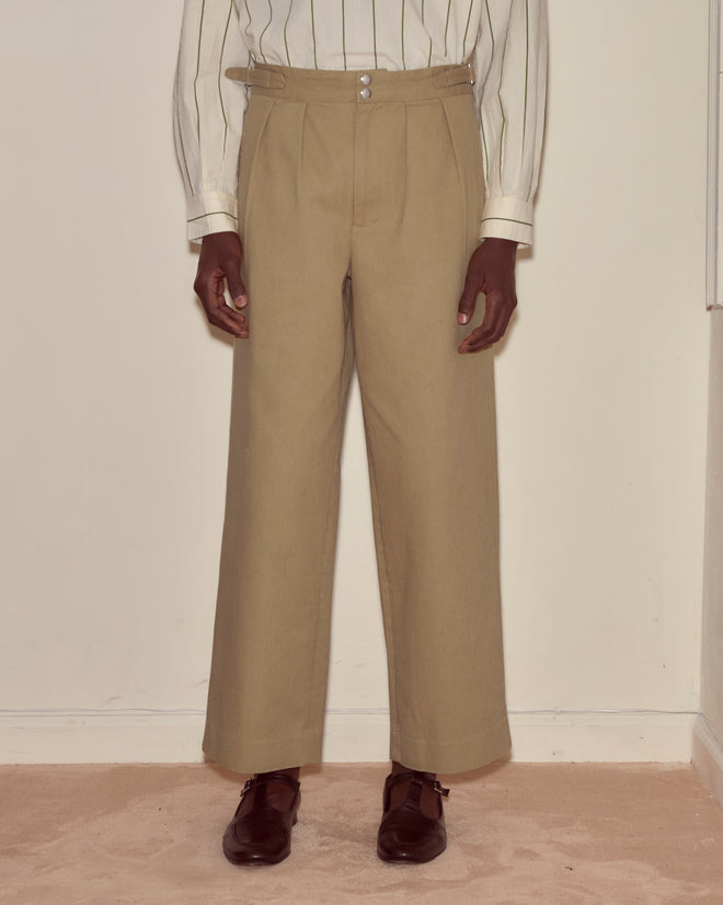 A person wearing Wide Leg Snap Trousers - Khaki, a white striped shirt, and brown shoes stands against a cream wall.