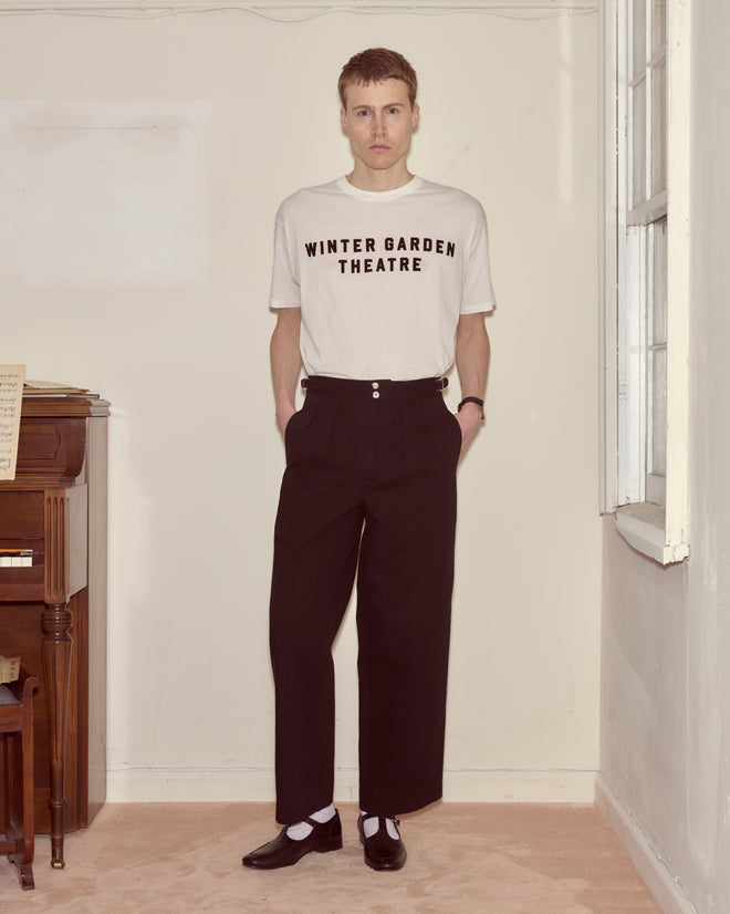 A person stands indoors by a piano, wearing a "Winter Garden Theatre" t-shirt and Wide Leg Snap Trousers - Black.