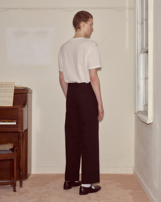 A person in a white t-shirt and Wide Leg Snap Trousers - Black stands facing a window in a plain room with a piano.