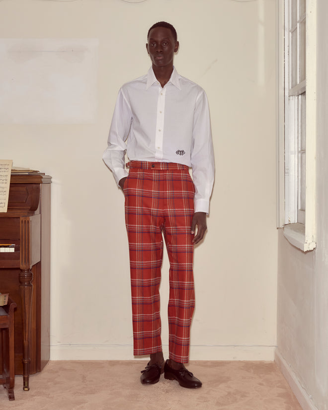Someone wearing a white shirt and Gable Plaid Trousers stands indoors beside a piano and a window.