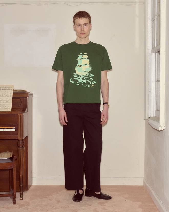 Indoors, a man stands by a piano wearing the Pirate Ship Tee—crafted from 100% cotton for comfort—inspired by Peter Pan. He pairs it with black pants and black shoes.