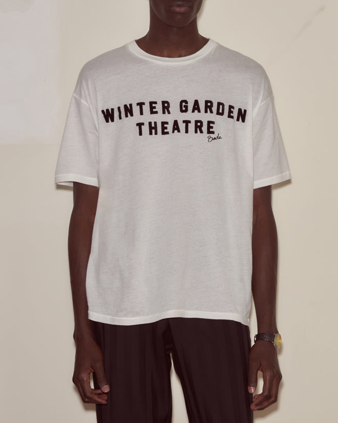 A person wears the "Theatre Tee," featuring a white design and black pants, standing against a plain background—a subtle, stylish tribute to Broadway and timeless classics like Peter Pan.