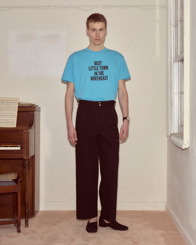 A man in a blue Laketown Tee that says "BEST LITTLE TOWN IN THE NORTHEAST" stands in a room by a piano.