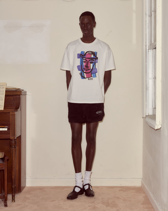 Indoors, a person wears the Chapeau Tee with abstract colorblocking, black shorts, white socks, and black shoes.