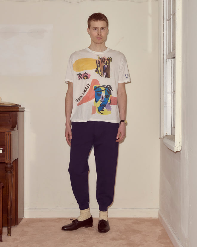 A person stands indoors by a piano wearing the Collage Tee, dark pants, socks, and black shoes.