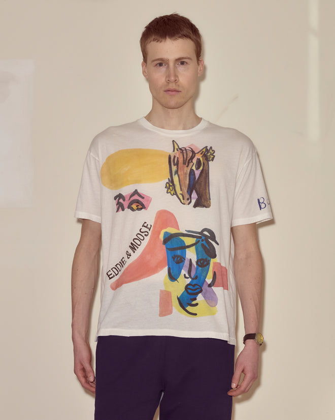 A man stands against a plain background, wearing the Collage Tee—a white souvenir shirt with abstract colorful faces and text, inspired by modernist art.