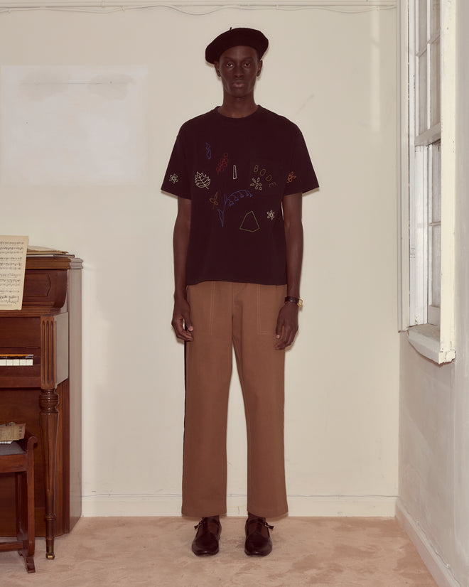 Wearing the Embroidered Field Guide Tee, a person in a beret, black shirt, and tan pants stands by a piano in a softly lit room.