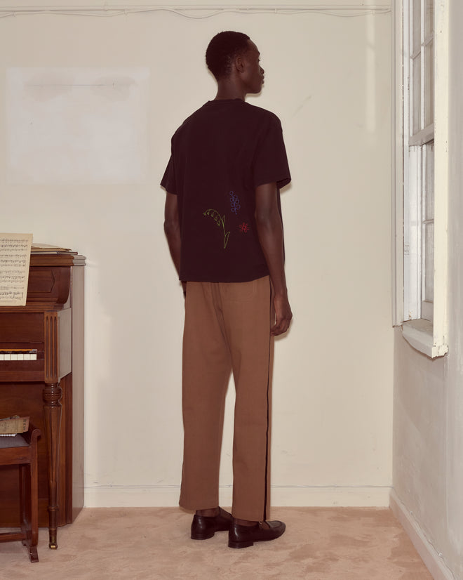 A person stands facing a wall in a room with a piano, wearing the Embroidered Field Guide Tee and brown pants.