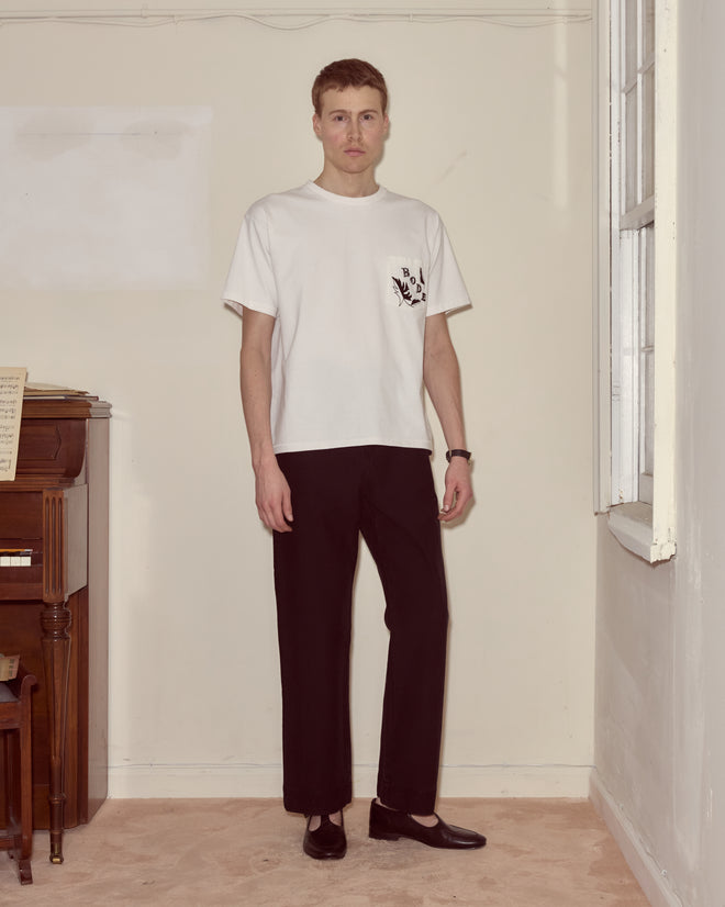 A person stands indoors by a piano and window, wearing a white Beaded Leaf Pocket Tee made of 100% cotton, black pants, and black shoes.