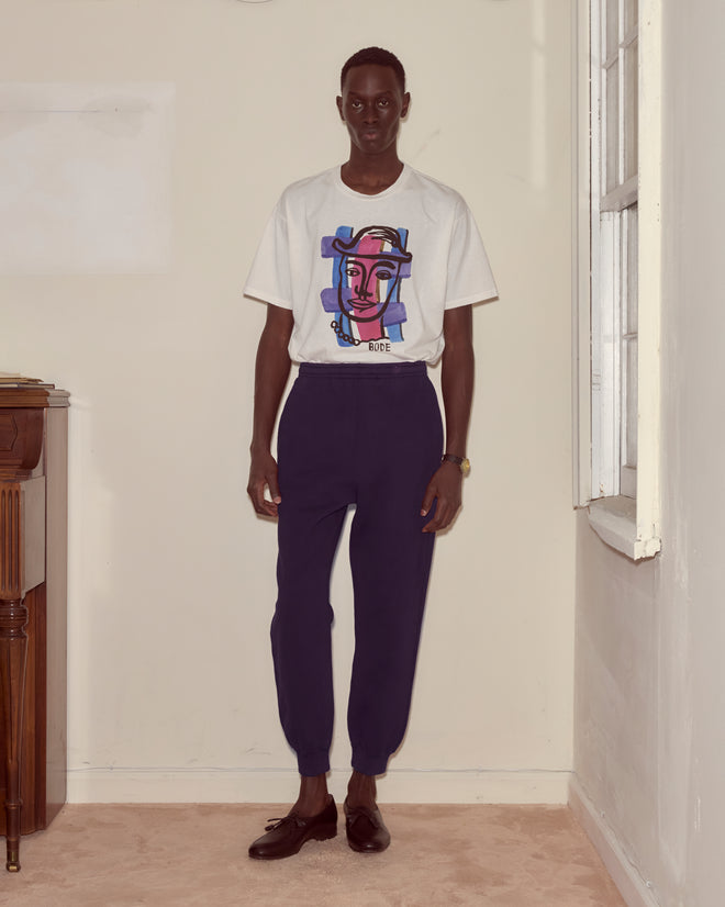 A man stands indoors near a window and piano, wearing a graphic t-shirt, dark purple joggers, and dark loafers.