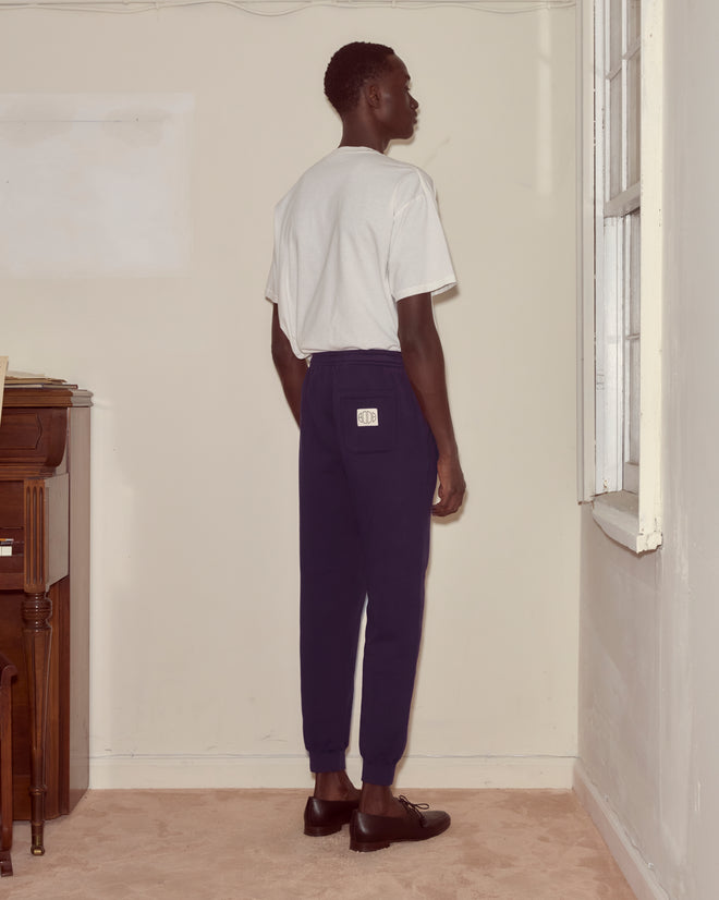 A man wearing a white t-shirt and dark purple Joggers stands facing a wall in a sparsely furnished room.